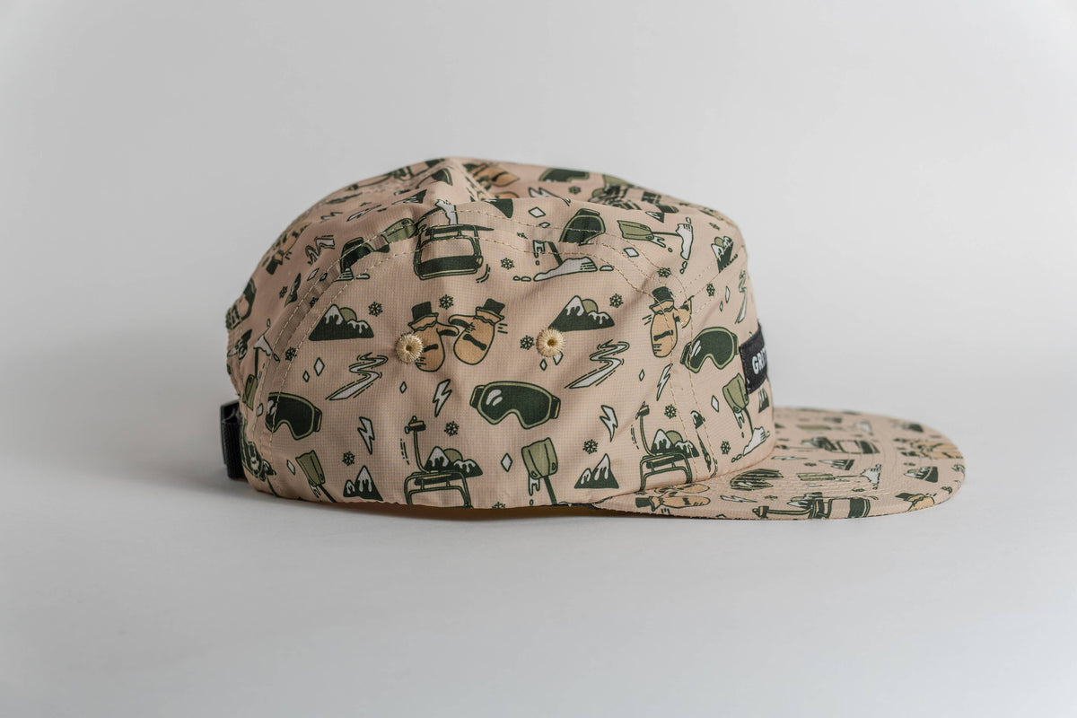 The First Chair 5-Panel Hat is a beige, flat-brim cap made from performance polyester, decorated with green and brown outdoor-themed illustrations (trees, mountains, backpacks, sunglasses) on a white background.