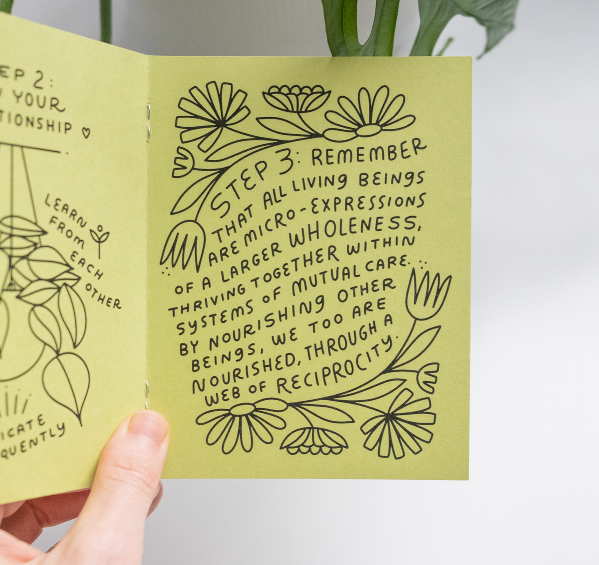 A hand holds an open "How to Keep A Plant Alive Zine" with hand-drawn flowers and handwritten notes on mutual care, nourishment, and reciprocity among all living beings as part of a greater wholeness.