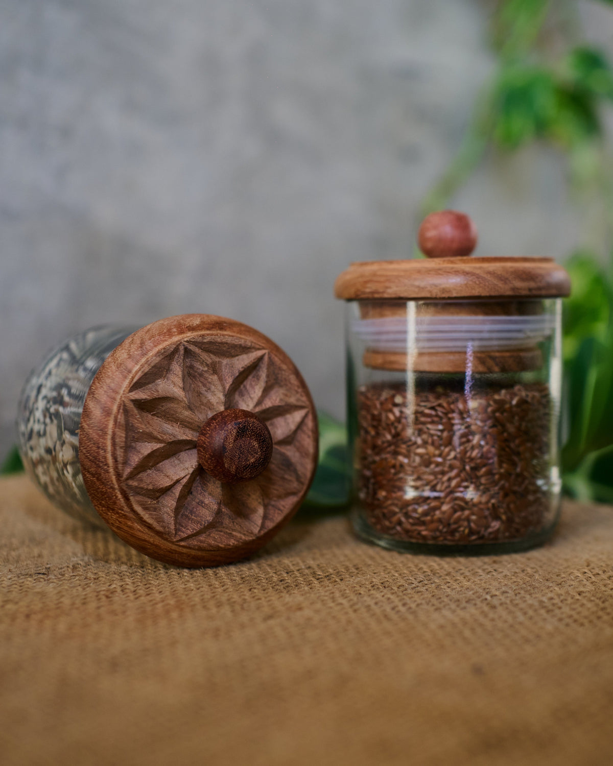 Two Karafuu Upcycled Glass Jars (Medium) with carved wooden lids—one upright and filled with brown flax seeds, one lying on its side—rest on burlap with green leaves, set against a gray background.