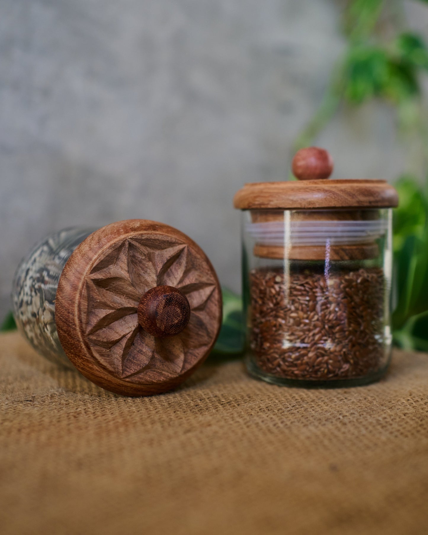 Two Karafuu Upcycled Glass Jars (Medium) with carved wooden lids—one upright and filled with brown flax seeds, one lying on its side—rest on burlap with green leaves, set against a gray background.