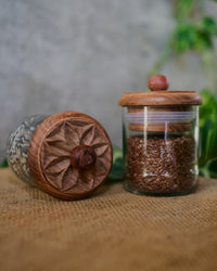Two Karafuu Upcycled Glass Jars (Medium) with carved wooden lids—one upright and filled with brown flax seeds, one lying on its side—rest on burlap with green leaves, set against a gray background.