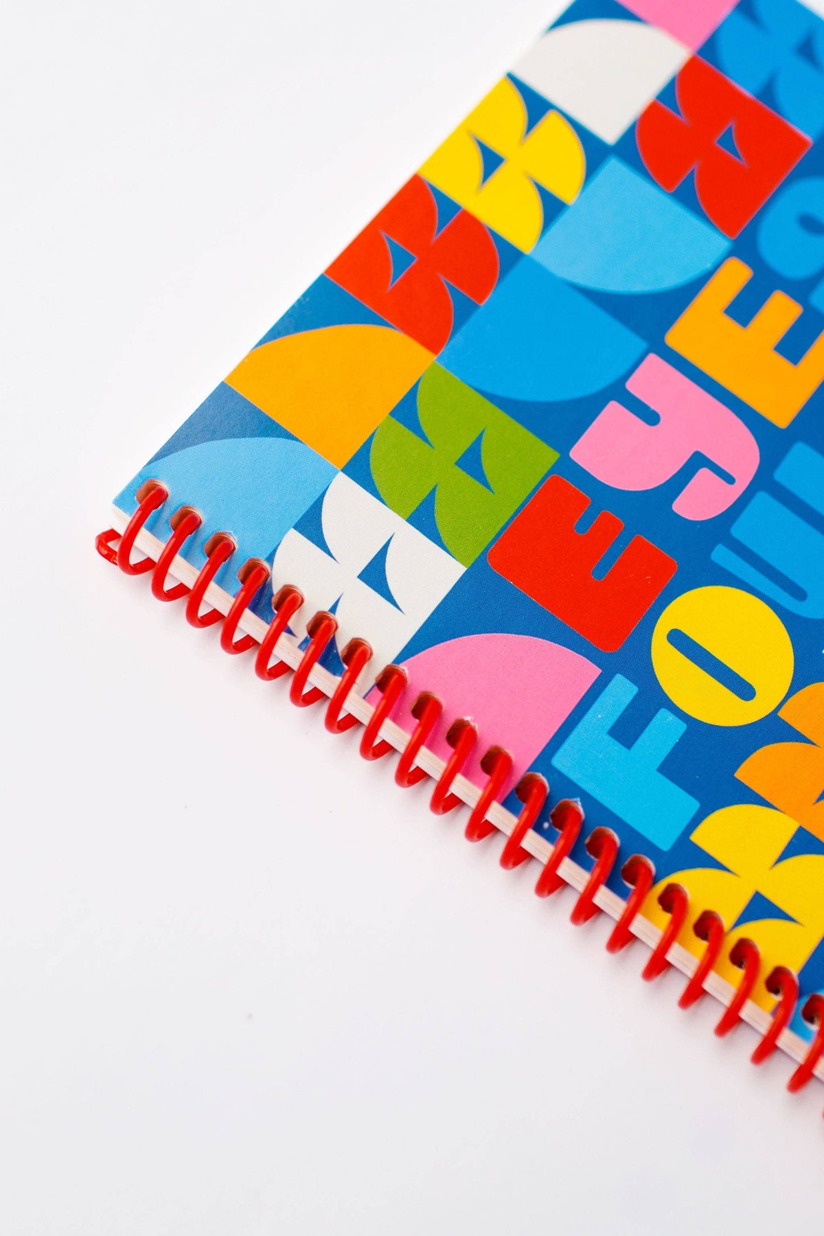 The Eyes Forward, Heart Open Journal is a vibrant spiral-bound notebook with a bold geometric and lettered cover design in blue, yellow, red, pink, and orange, finished with a striking red spiral. Perfect for capturing creative ideas or notes.