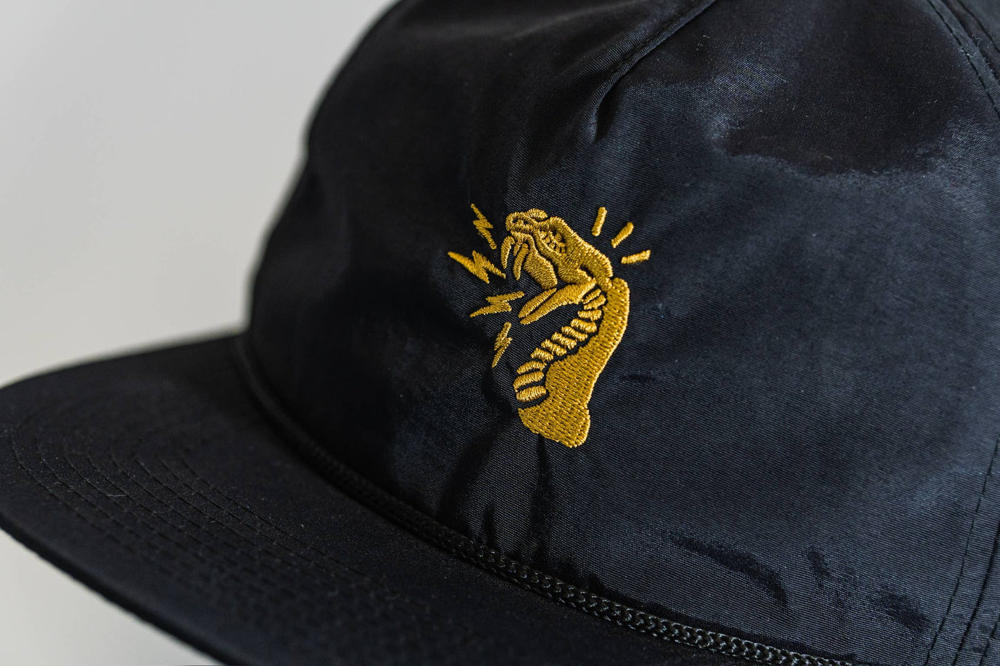 A close-up of the Fearless 5-Panel Hat by Gritty Habits, black with a gold embroidered coiled snake, mouth open and lightning bolts encircling its head.