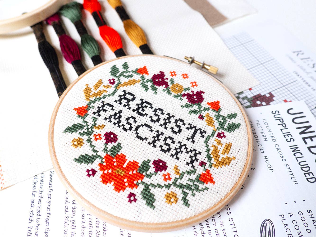 The Resist Fascism Cross Stitch Kit features a round hoop holding fabric stitched with "RESIST FASCISM," surrounded by colorful flowers and leaves. This beginner-friendly kit is handmade in Portland and includes threads and a pattern sheet.