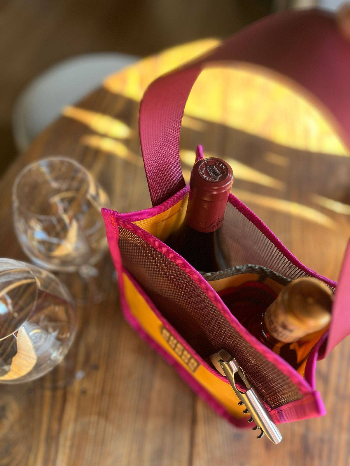 The Two-Bottle-Bag | Cava on the Playa, a vibrant tote for two wine bottles and a corkscrew, rests on a wooden table beside empty glasses as sunlight streams in—ideal for your next picnic or gathering.
