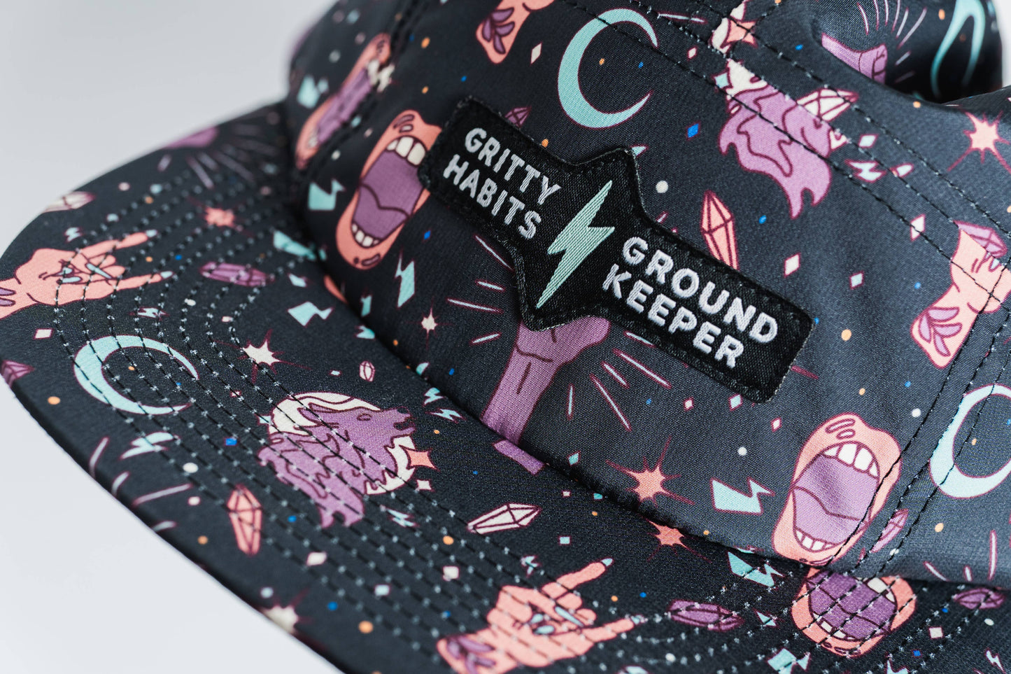 A close-up of the Gritty Habits x Ground Keeper: Howl Collection 5-Panel Hat, a black women-owned cap with playful moons, hands, mouths, and lightning bolts. The front patch reads GRITTY HABITS | GROUND KEEPER separated by a blue lightning bolt.