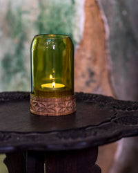 The Upepo Tea Light Holder, crafted from an upcycled wine bottle with a wooden base, glows on a dark ornate wooden table against a blurred, textured wall background.