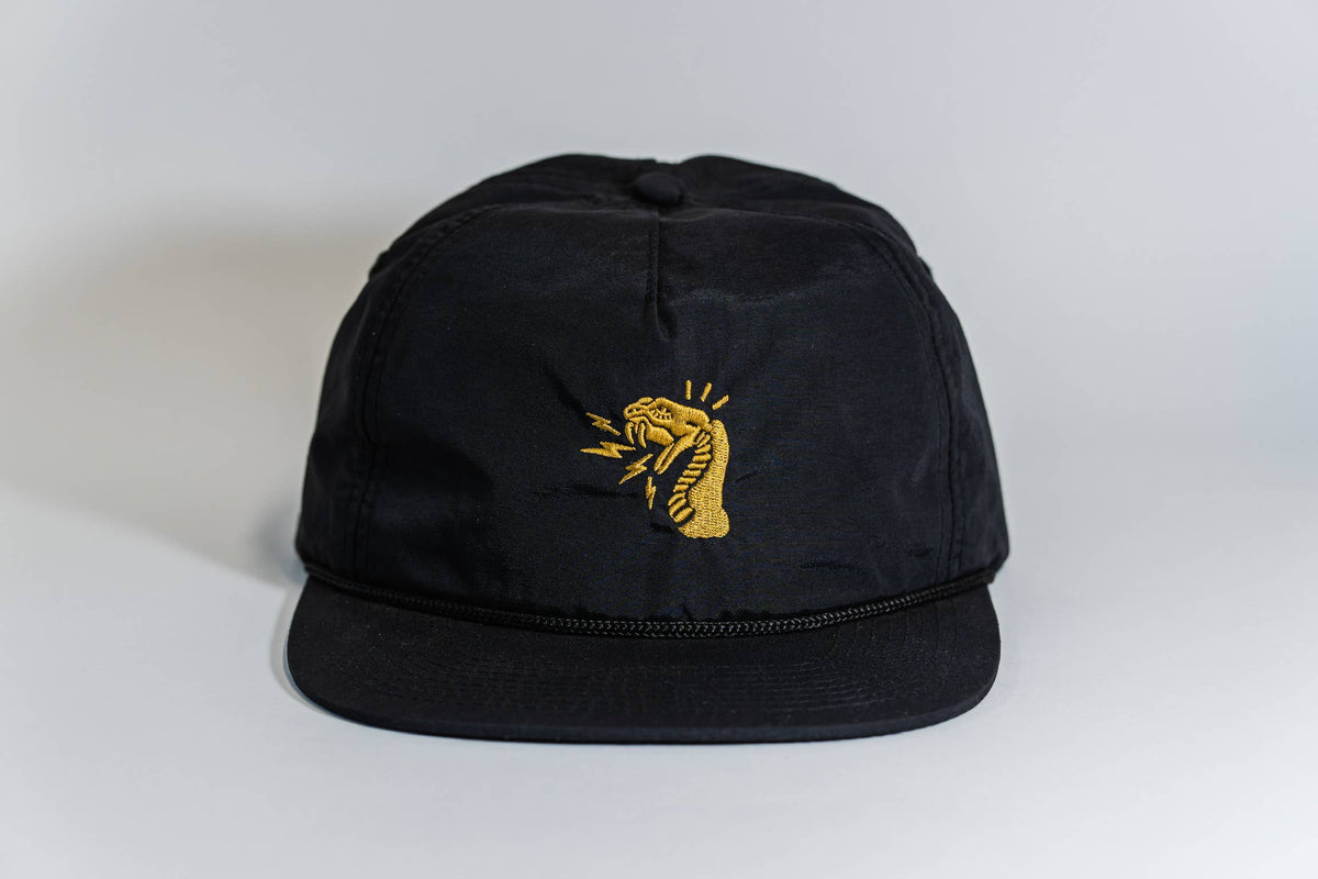 The Fearless 5-Panel Hat is a black cap with an embroidered yellow cobra, mouth open and tongue out, on the front. Displayed on a white background, it's part of the Gritty Habits collection.