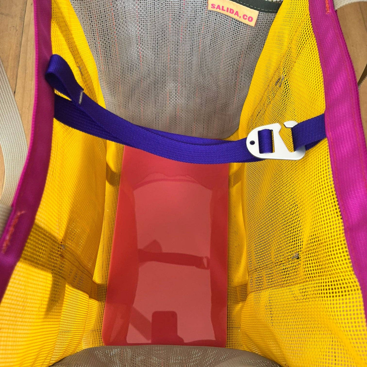 The Mother | Home Base is a mesh tote with yellow sides, purple trim, and a blue strap. Inside, it has a pink rectangular acrylic base and a SALIDA CO label on the inner rim, showcasing Buena Goods' signature style.