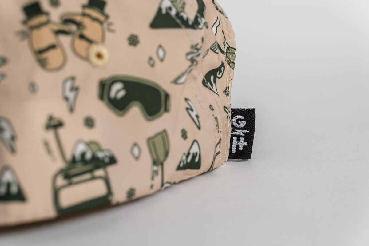 Close-up of the beige First Chair 5-Panel Hat made from performance polyester, featuring green and yellow winter-themed prints like ski goggles, mittens, and mountains. A small black tag with a white logo is sewn into its water-resistant edge.