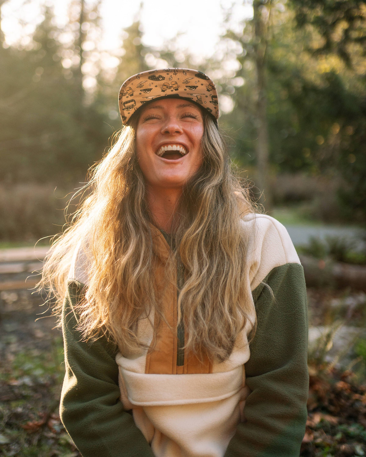 A woman with long wavy hair, wearing a patterned First Chair 5-Panel Hat and a fleece jacket, smiles and laughs while sitting outdoors in a sunlit wooded area.