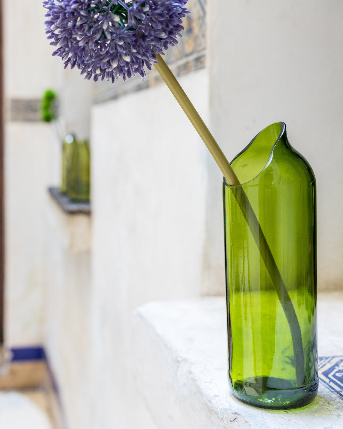 The Kikuba Upcycled Vase in green holds a long-stemmed purple allium on a white indoor ledge. Another green vase is blurred in the background—a stylish, sustainable accent for any home.