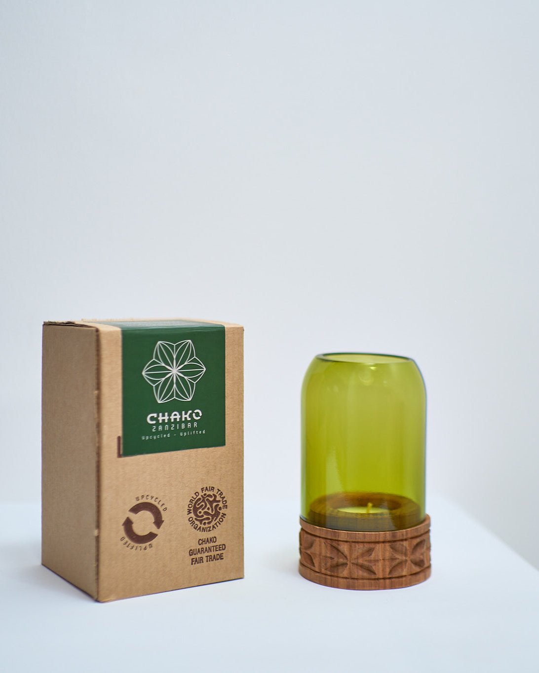 A cardboard box labeled CHAKO sits beside the Upepo Tea Light Holder, a fair trade Zanzibar candle holder with a green glass cover and carved wooden base, all arranged on a white surface against a plain backdrop.