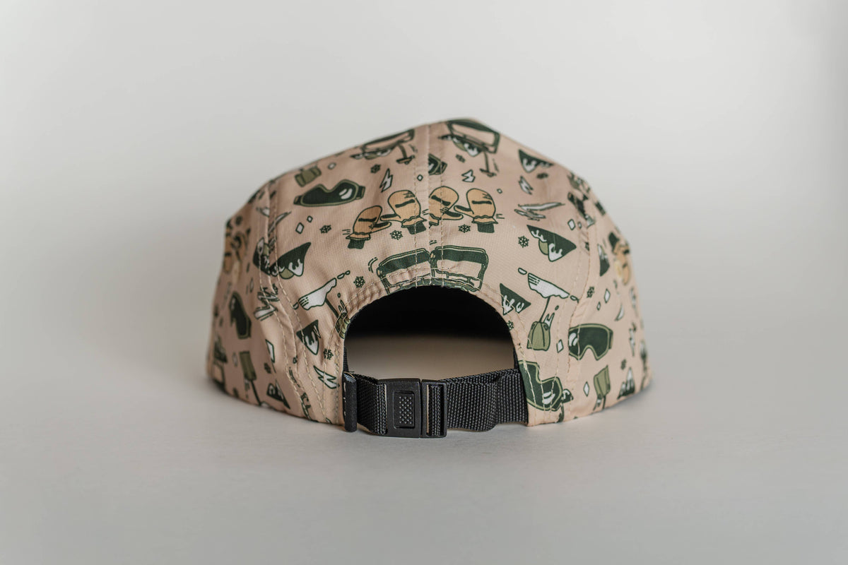The First Chair 5-Panel Hat in beige features a black adjustable strap and green and yellow bottle, glass, and hat illustrations. Made from water-resistant performance polyester for style and durability.