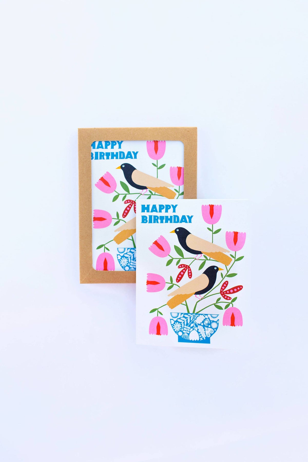 The Happy Birthday Nordic Birds Greeting Card features two black and yellow birds on a floral branch in a blue vase, surrounded by pink and red flowers. The FSC-certified card displays "HAPPY BIRTHDAY" in blue letters, with another card partially visible behind.
