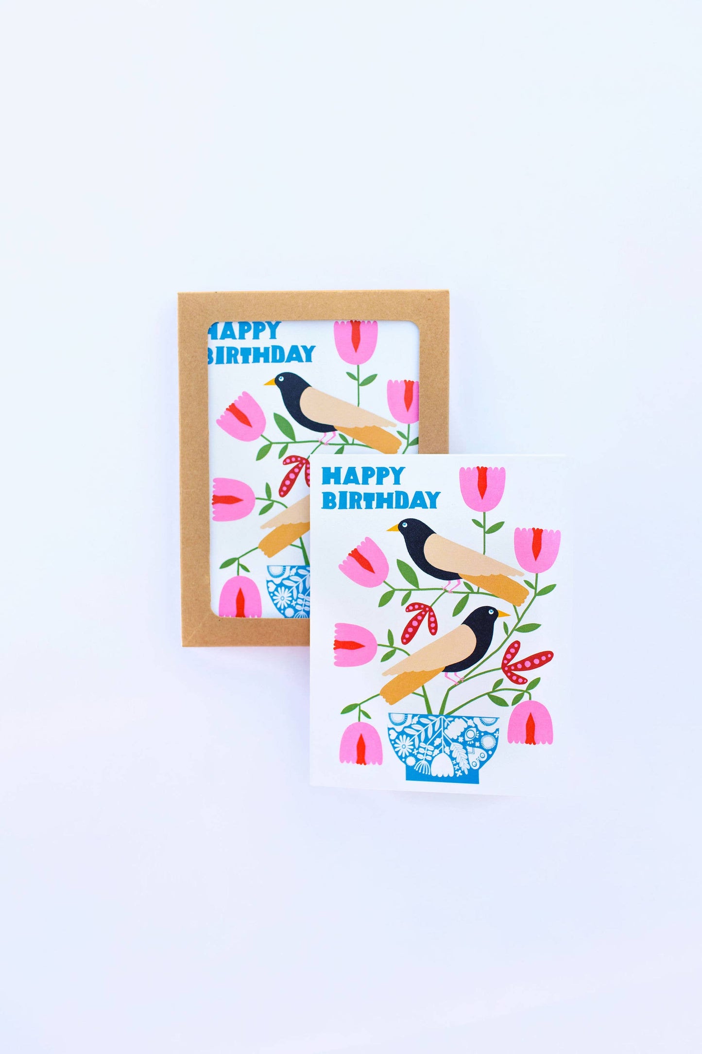 The Happy Birthday Nordic Birds Greeting Card features two black and yellow birds on a floral branch in a blue vase, surrounded by pink and red flowers. The FSC-certified card displays "HAPPY BIRTHDAY" in blue letters, with another card partially visible behind.