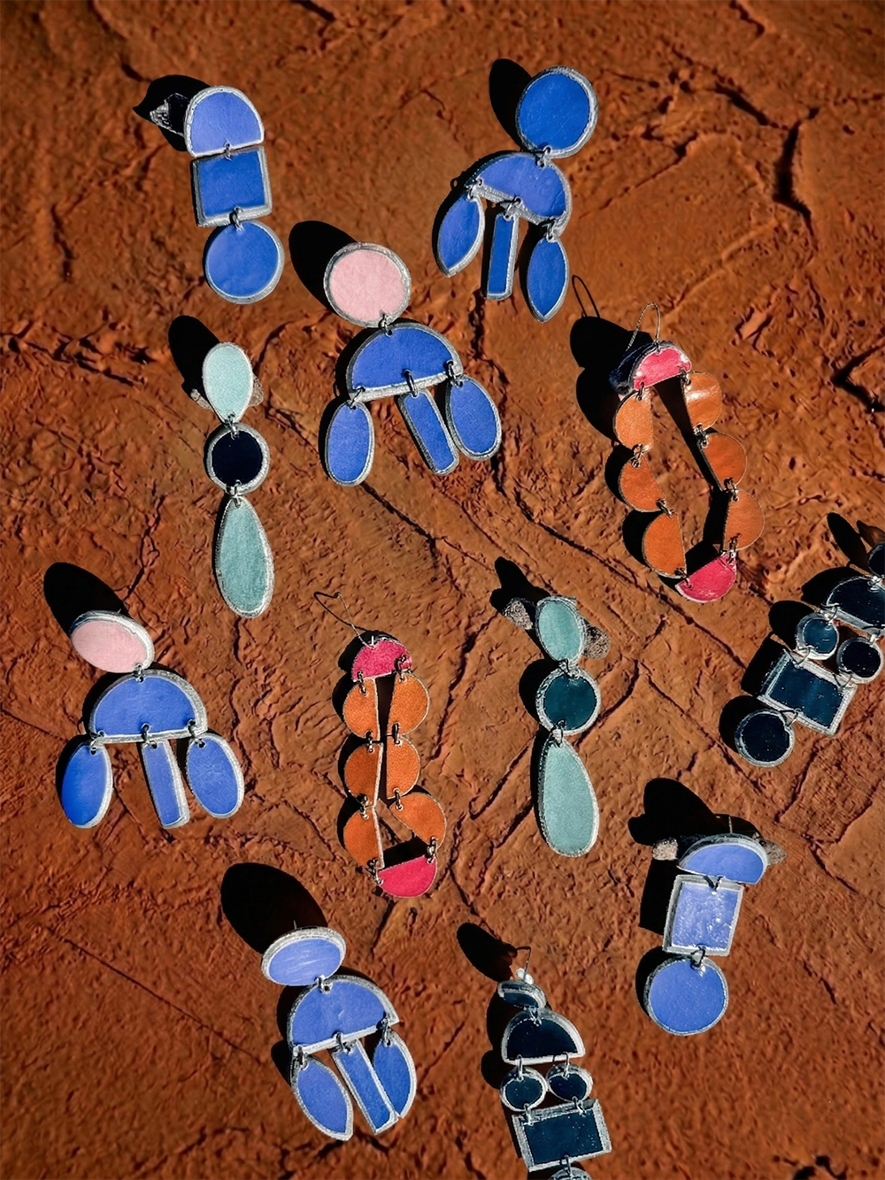 Sisanda Paper Pulp Earrings by Quazi Design, in circular, oval, and rectangular shapes and blue, green, orange, and pink colors, are displayed on a textured reddish-brown surface.