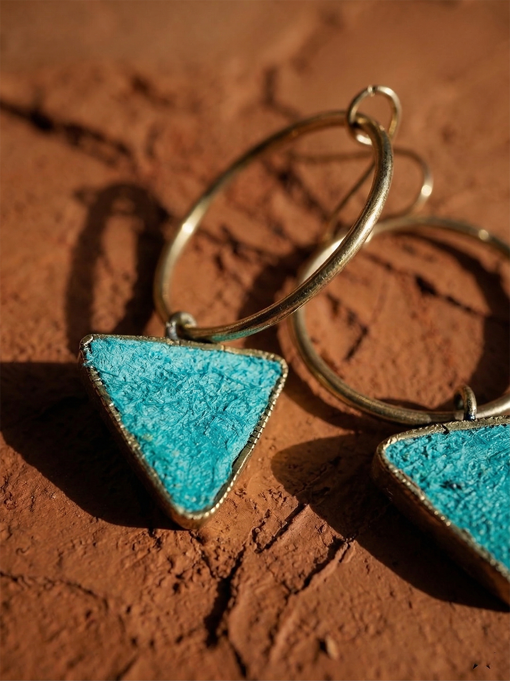 Close-up of Era Paper Pulp Earrings featuring handcrafted, textured, turquoise-colored triangle pendants displayed on a rustic brown surface.