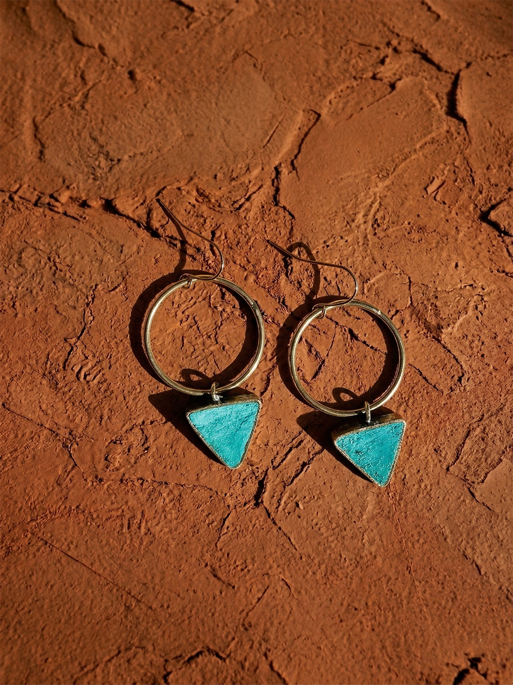 The Era Paper Pulp Earrings, a pair of silver hoops with turquoise triangle pendants, rest on a textured, reddish-brown surface, casting subtle shadows.