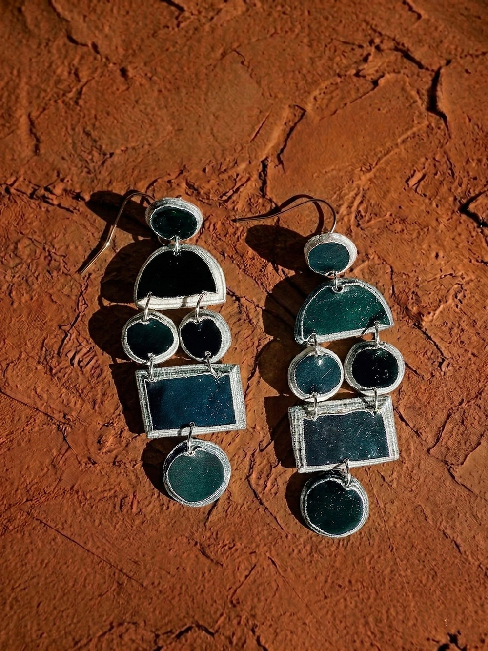Nokuthula Paper Pulp Earrings by Quazi Designs: geometric silver earrings handcrafted from recycled newspaper with black circular, semicircular, and rectangular shapes, shown on a textured reddish-brown surface in sunlight.