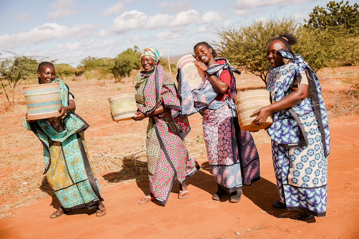 Four women dressed in vibrant patterned clothing stand outdoors on reddish soil, smiling and holding Artisan Safari woven baskets. Bushes and trees can be seen in the background beneath a partly cloudy sky.