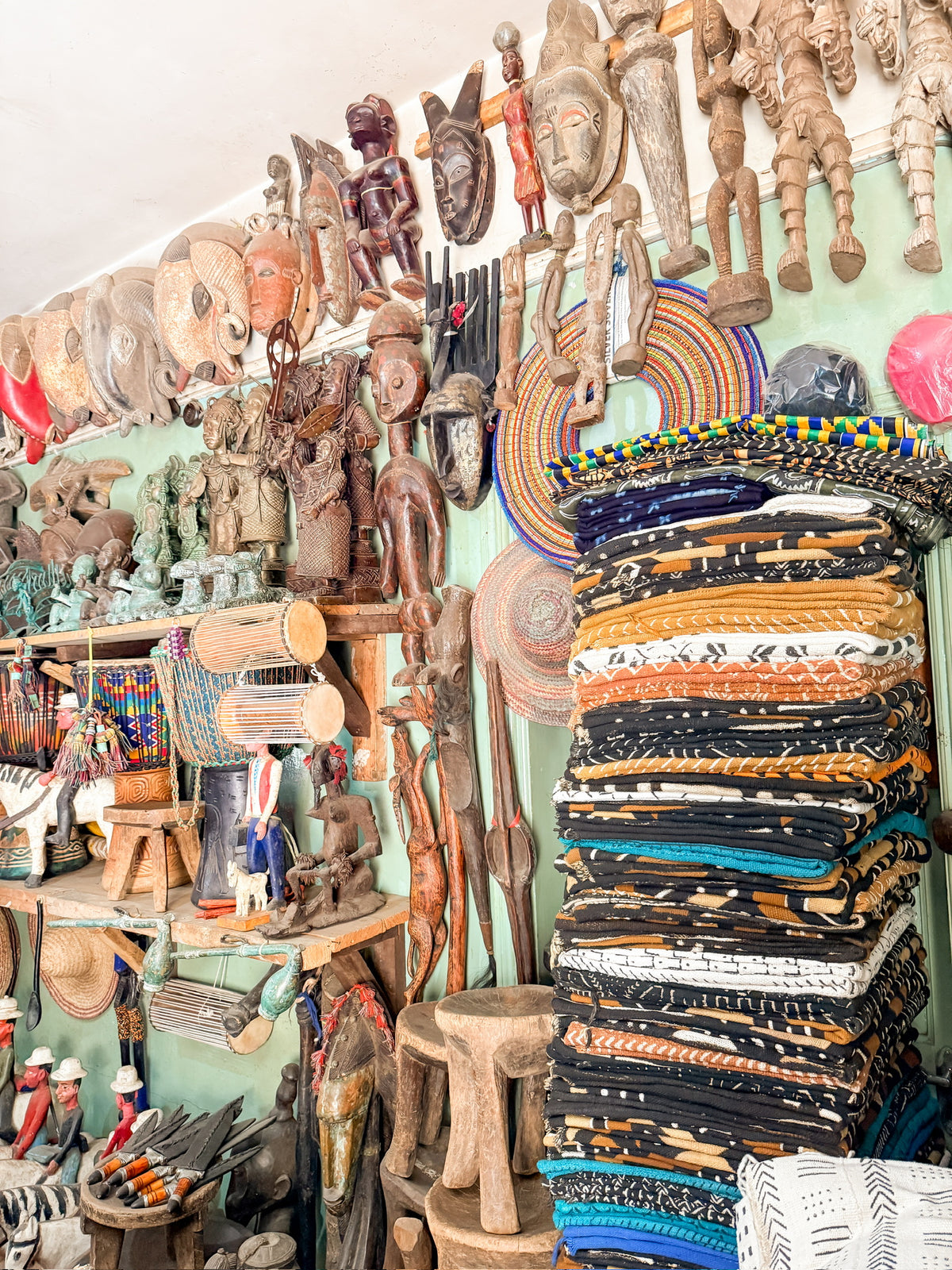 Inside Artisan Safari, colorful African textiles are stacked beside shelves filled with traditional wooden masks, sculptures, woven baskets, and other handcrafted artifacts in a vibrant, well-lit shop.