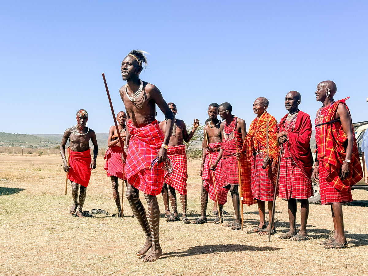 A group of people in traditional Maasai attire stand on dry grass under a clear blue sky, part of the vibrant scene captured by Artisan Safari. One person is jumping while others watch, all dressed in red and orange shukas.