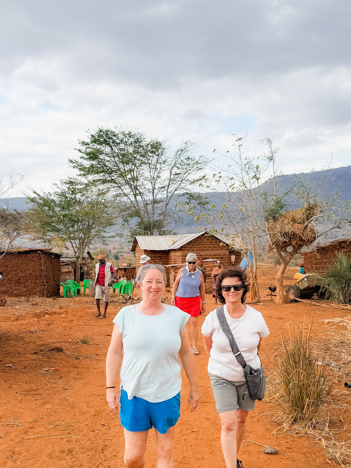 Four people walk along a dirt path in a rural area with small brick houses and dry vegetation, embodying the spirit of “Artisan Safari.” In the foreground, two women smile under a cloudy sky, with rolling hills visible in the distance.