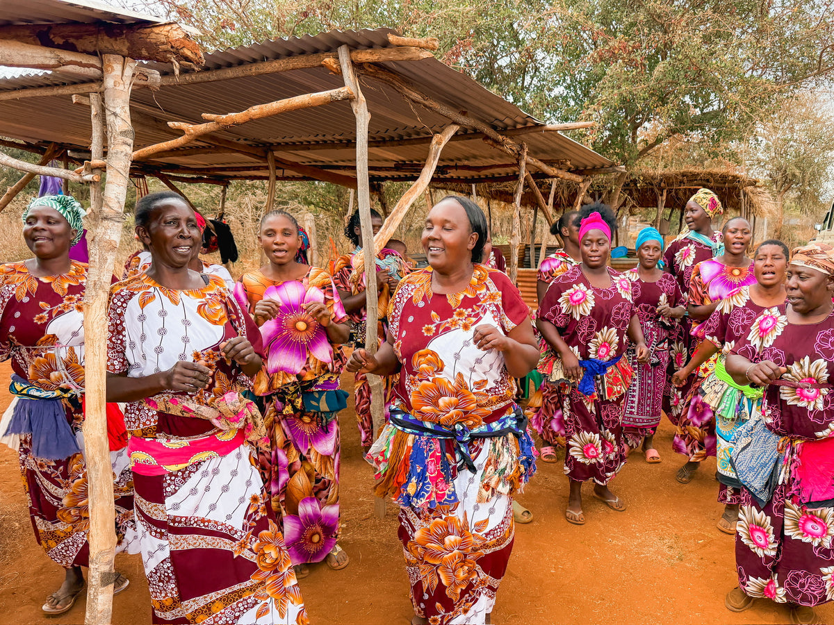 A group of women in colorful Artisan Safari dresses dance and sing outdoors under wooden shelters, surrounded by trees and reddish soil.