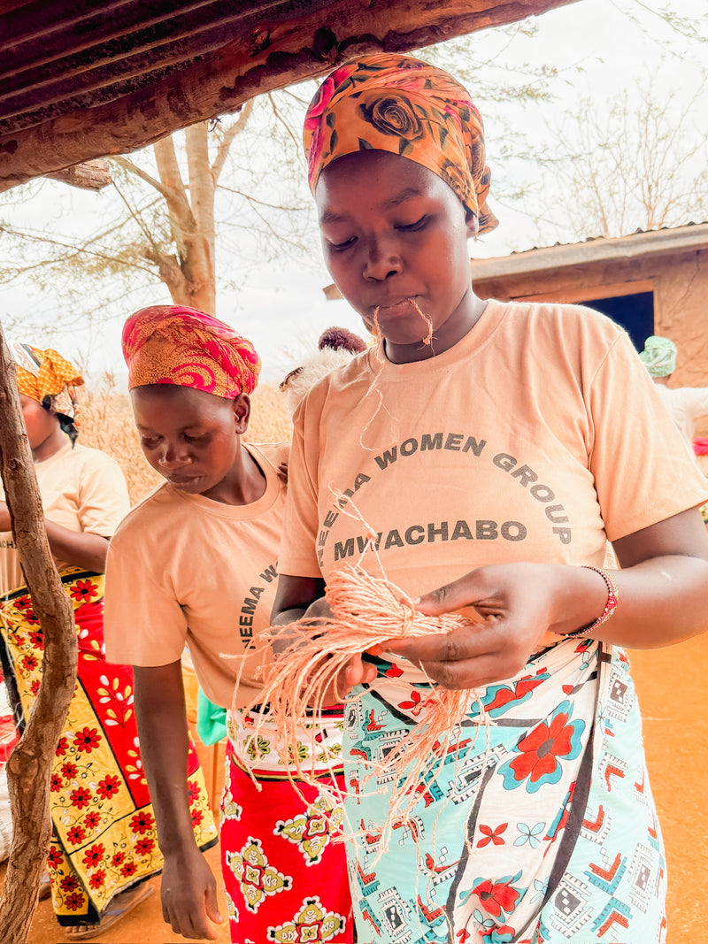 Two women wearing vibrant headscarves and patterned skirts weave fibers together outdoors as part of Artisan Safari. They wear matching shirts that read “HEEMA WOMEN GROUP MWACHABO.” One woman is focused intently on her weaving, with a building and a tree seen in the background.