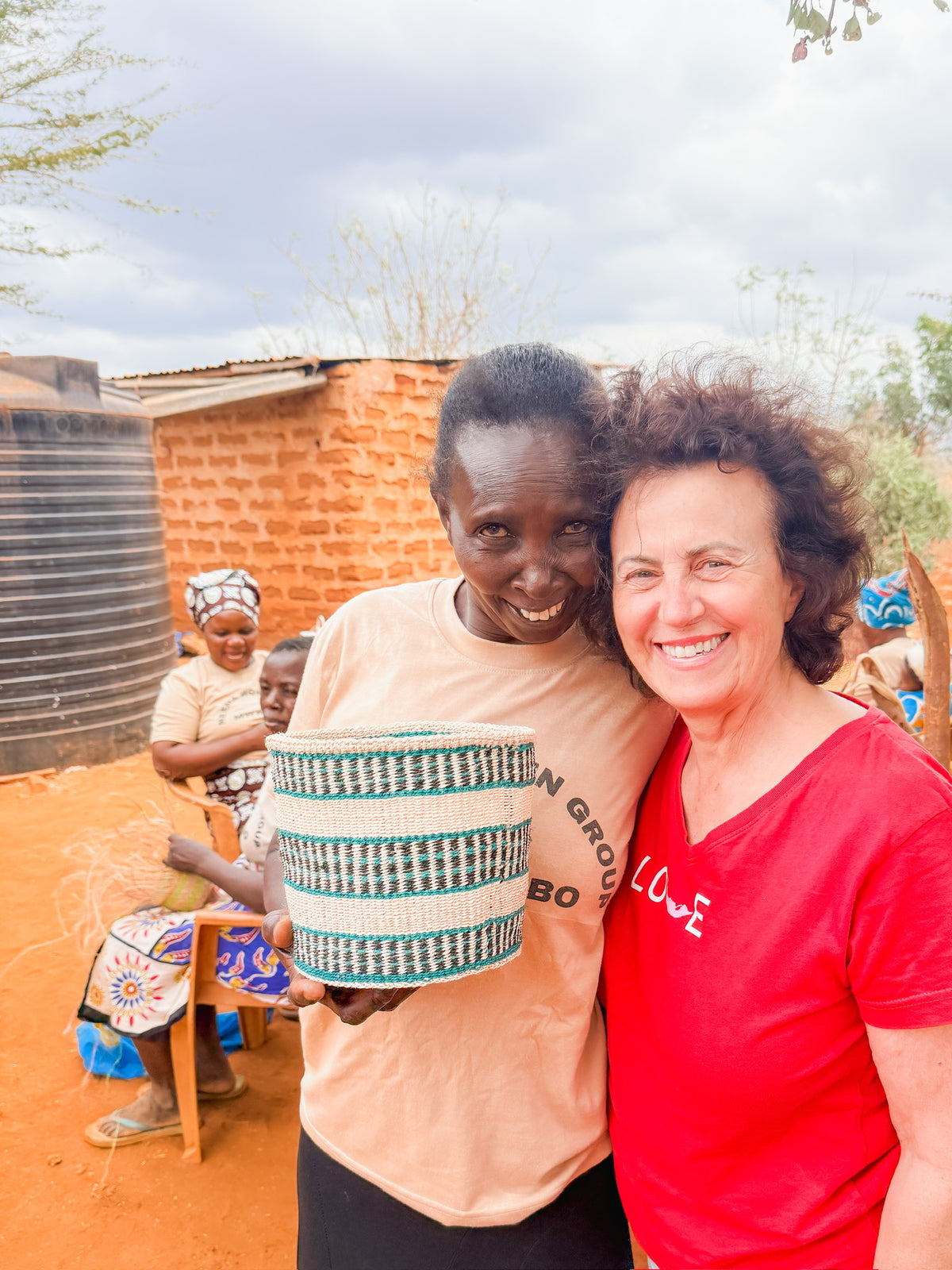 Four women from Artisan Safari smile and weave outside near a brick building and a large water tank. Two are seated, working on their craft, while one in the foreground holds a woven basket. The atmosphere is warm and cheerful.