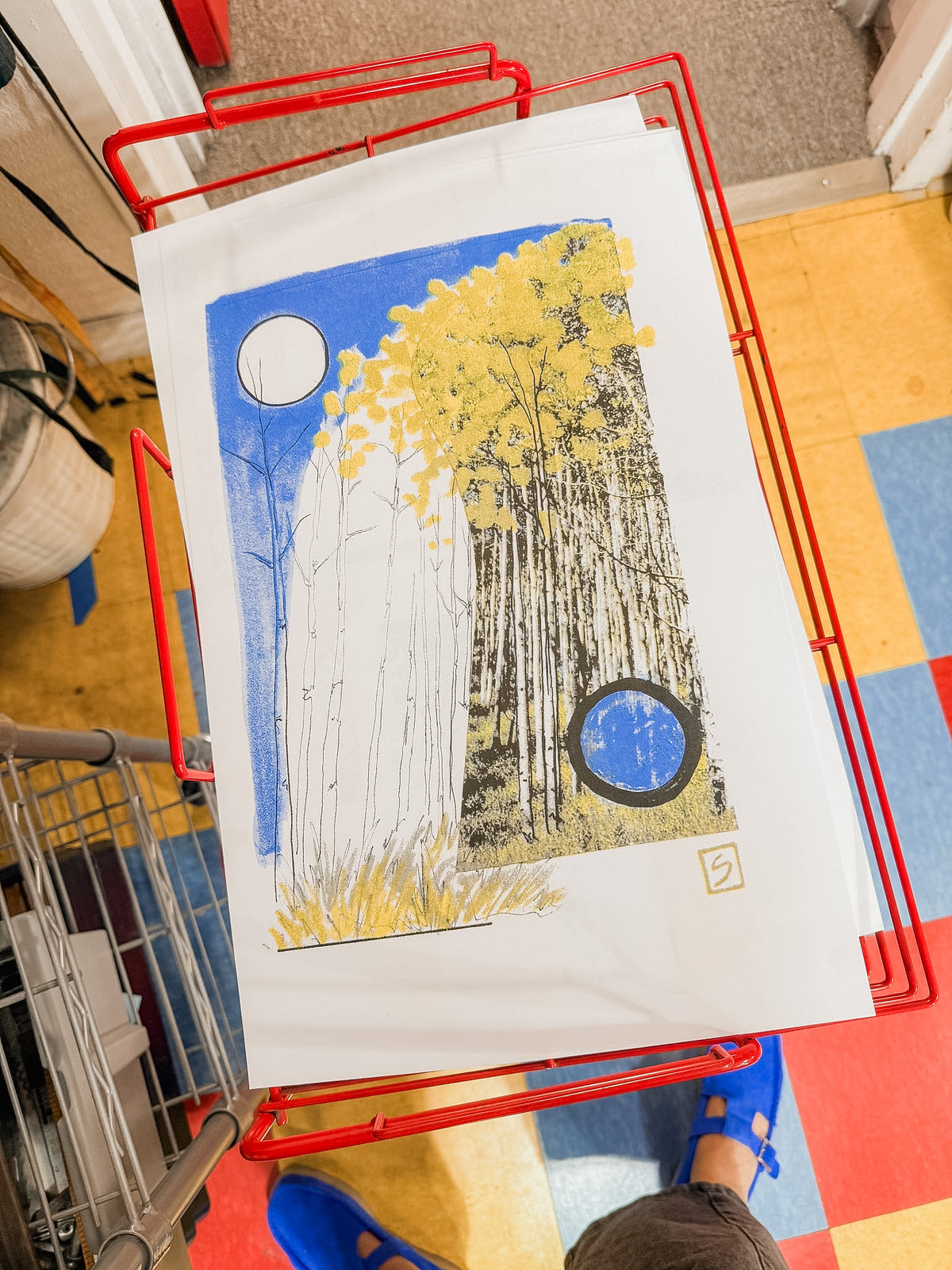 A vibrant limited edition “The Long Way | Riso Print,” featuring yellow trees and two circles, rests on white paper inside a red wire basket. The background reveals a tiled floor with red, blue, and yellow squares, with someone in blue shoes standing nearby.