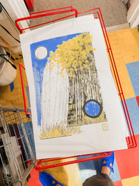 A vibrant limited edition “The Long Way | Riso Print,” featuring yellow trees and two circles, rests on white paper inside a red wire basket. The background reveals a tiled floor with red, blue, and yellow squares, with someone in blue shoes standing nearby.