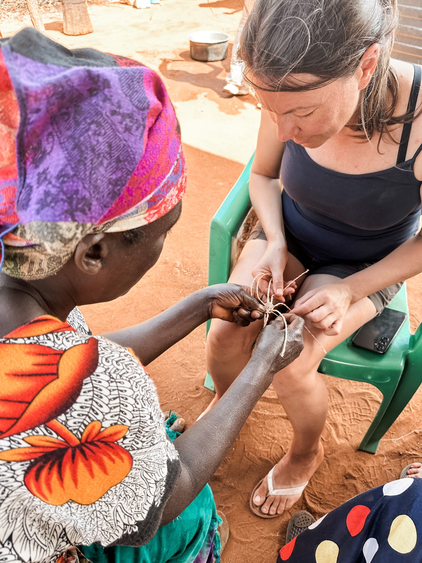 A woman in colorful clothes teaches another, seated on a green chair, to weave with natural fibers outdoors on reddish-brown soil during the Cycling Safari experience.