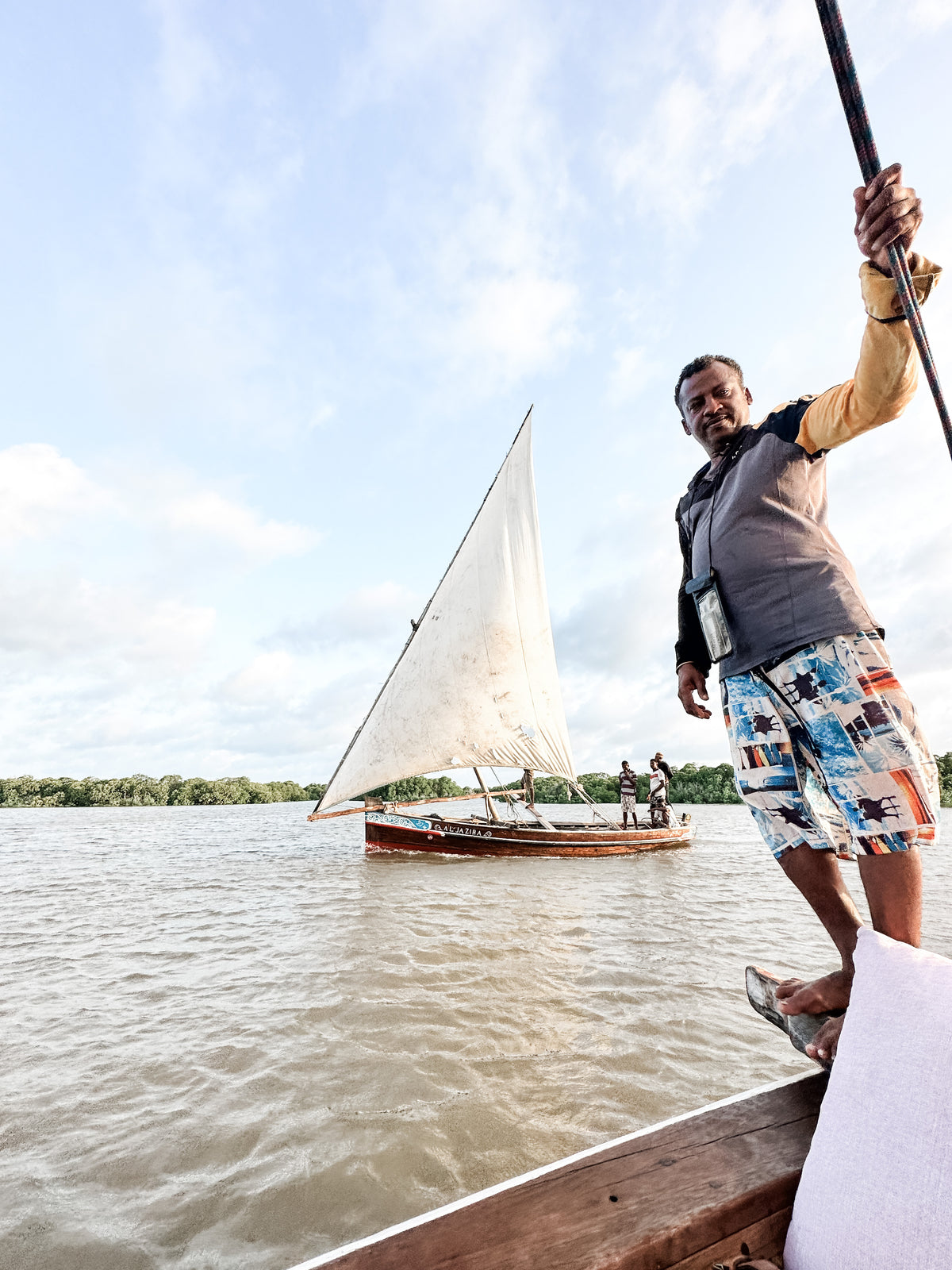A man stands on the Artisan Safari wooden boat holding a pole, with another sailboat and two people in the background on a wide river under a partly cloudy sky.