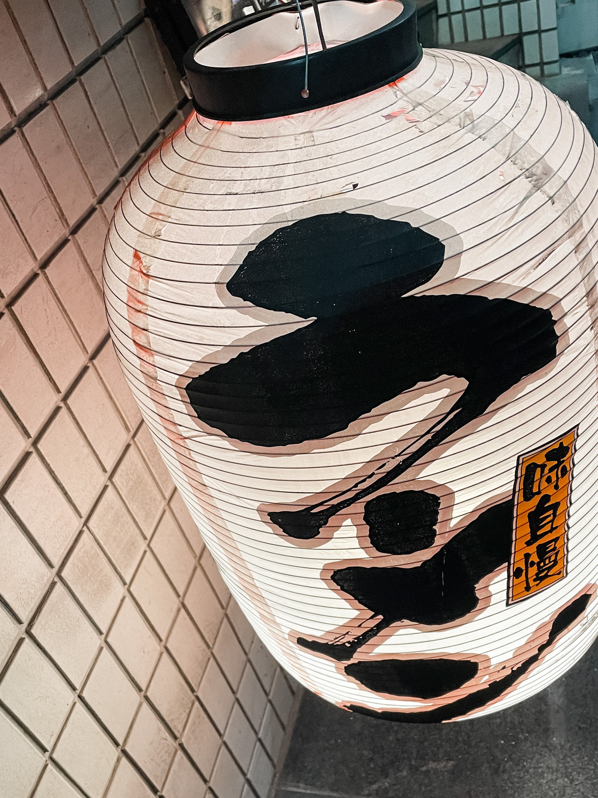 A Japanese Mystery Box with bold black kanji and a yellow label hangs against a tiled wall, casting soft light—perfect as a unique gift or travel souvenir for anyone who loves authentic Japanese decor.