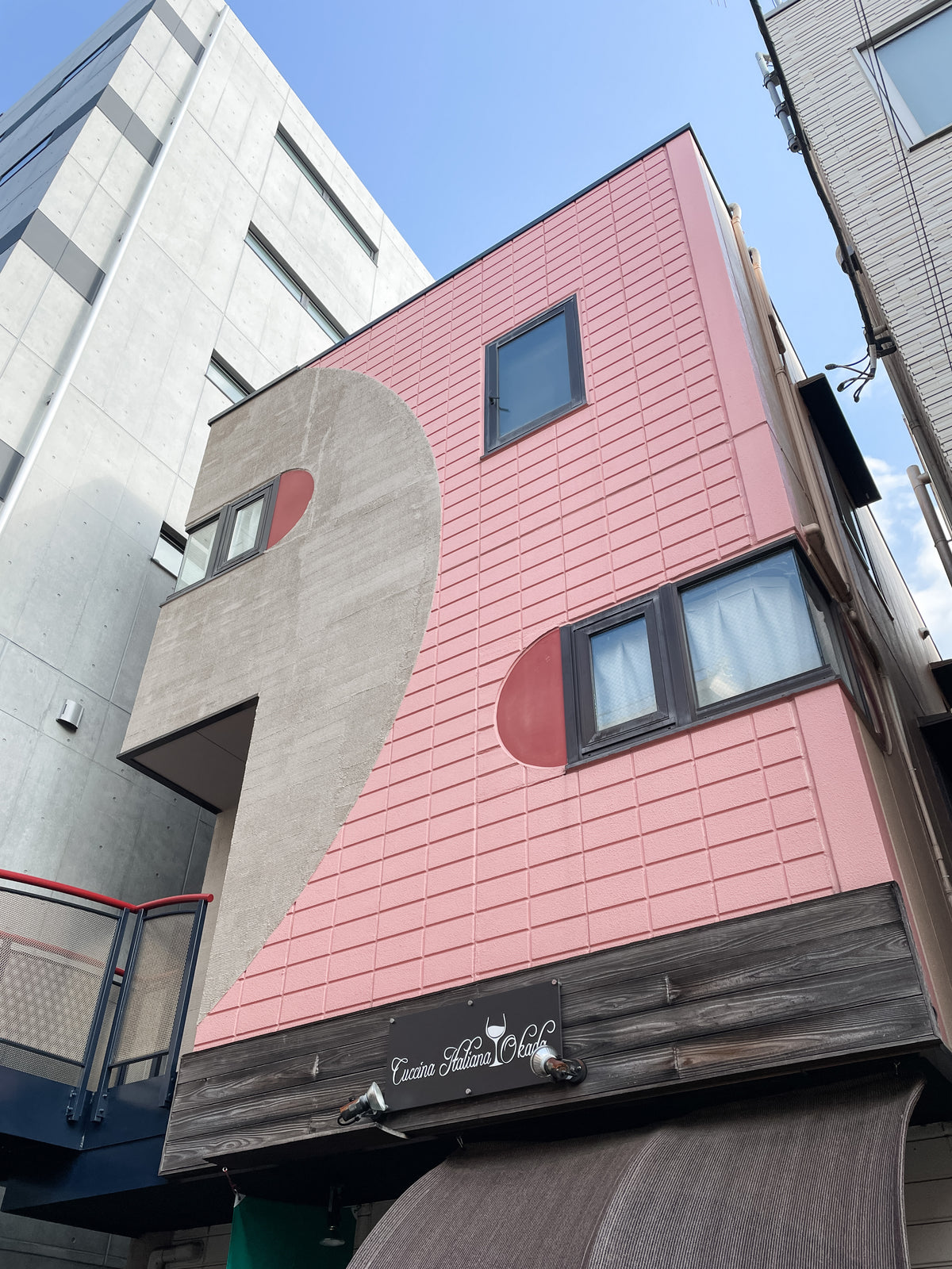 A modern building with a pink and beige facade, large stylized comma design, and a sign for Cursive Hiroo Cafe suggests Japanese Mystery Box gifts and travel souvenirs inside. The sky above is clear and blue.
