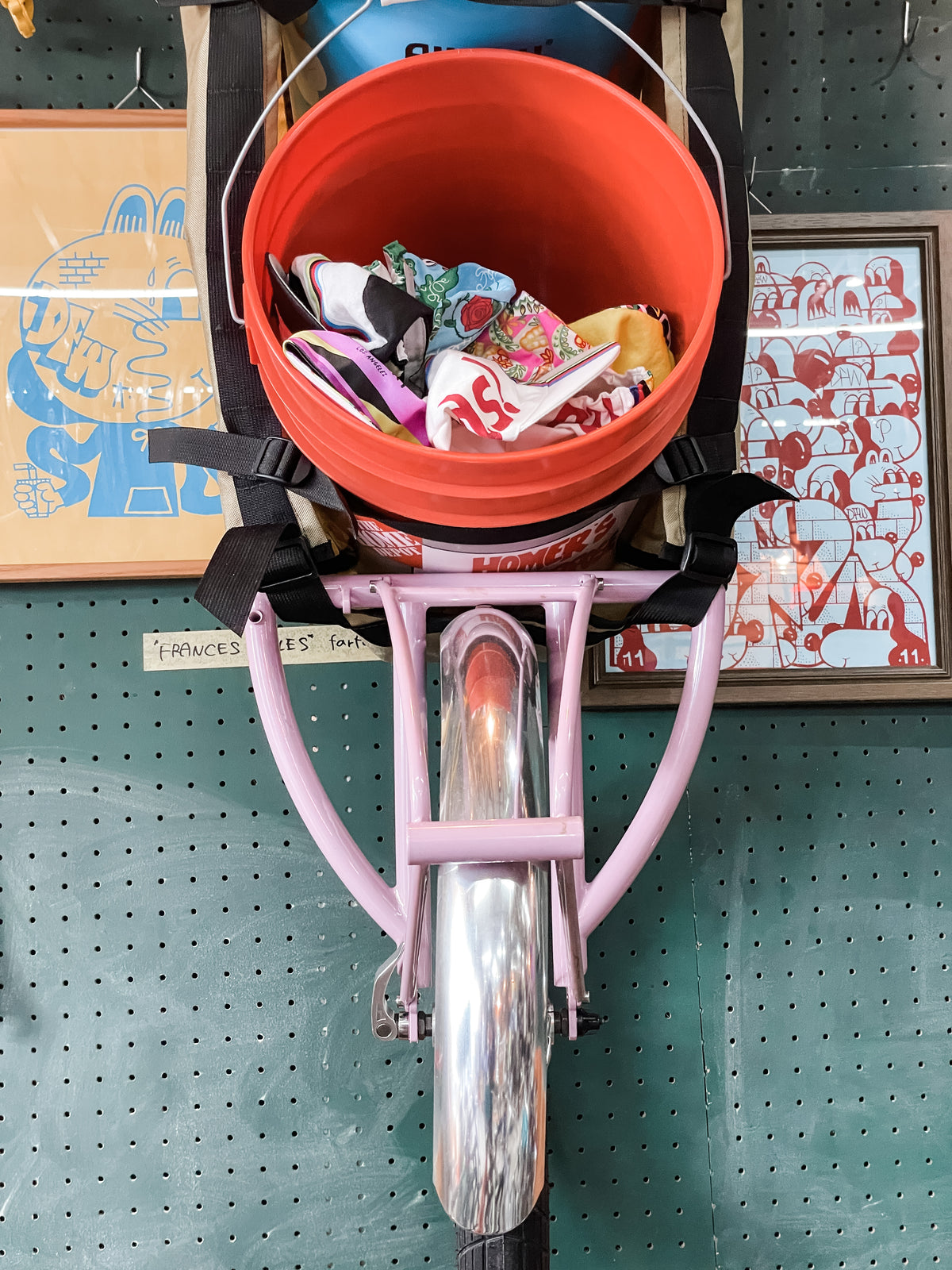 A pink unicycle is mounted vertically on a green pegboard wall. A red bucket holding a Japanese Mystery Box sits on the unicycle, with framed artwork visible in the background.