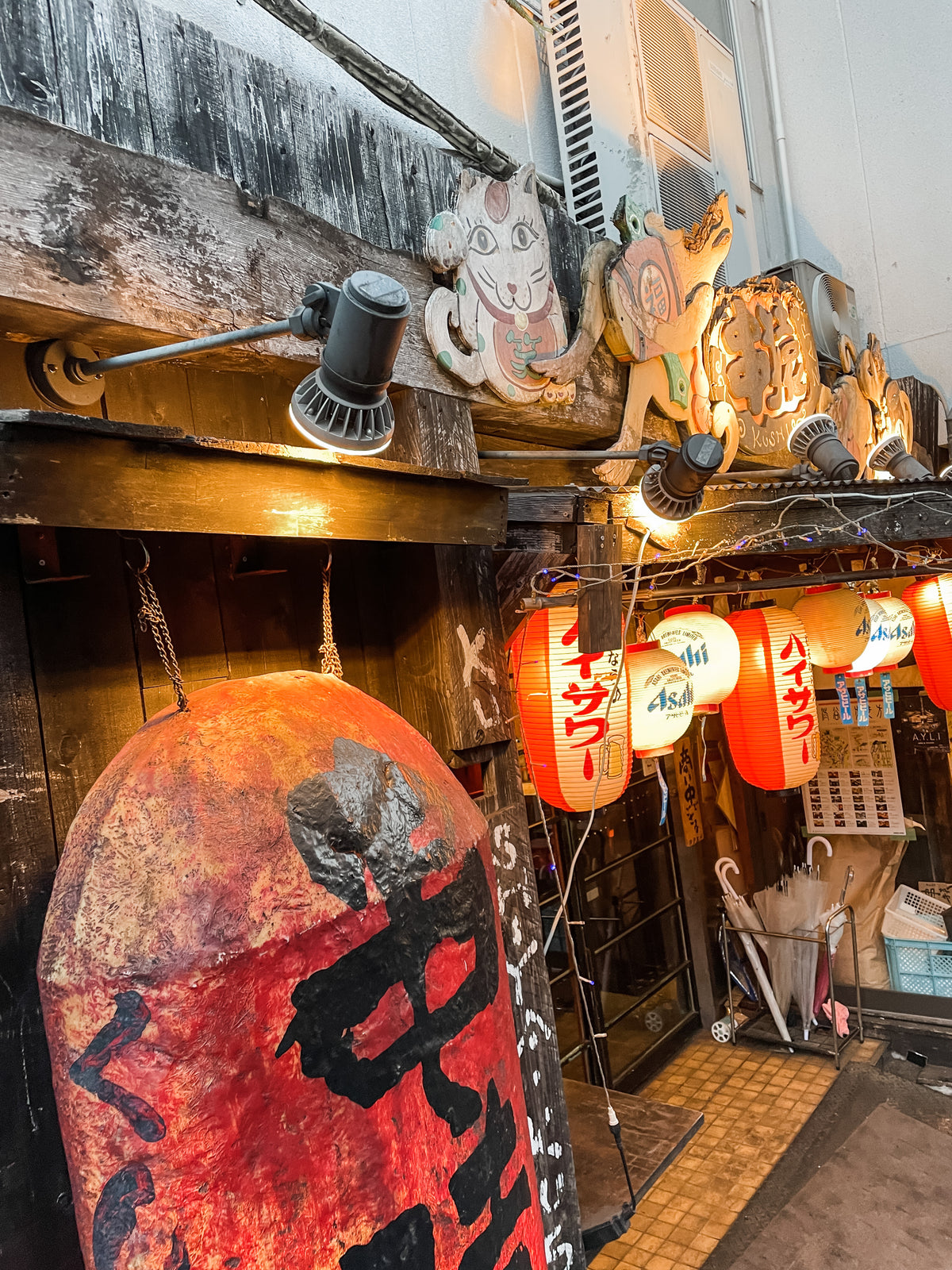 A cozy Japanese izakaya entrance with red lanterns, wooden signs, and animal-shaped decorations welcomes you. A large kanji lantern and umbrellas by the door hint at the surprises inside each Japanese Mystery Box—unique gifts or travel souvenirs await!.