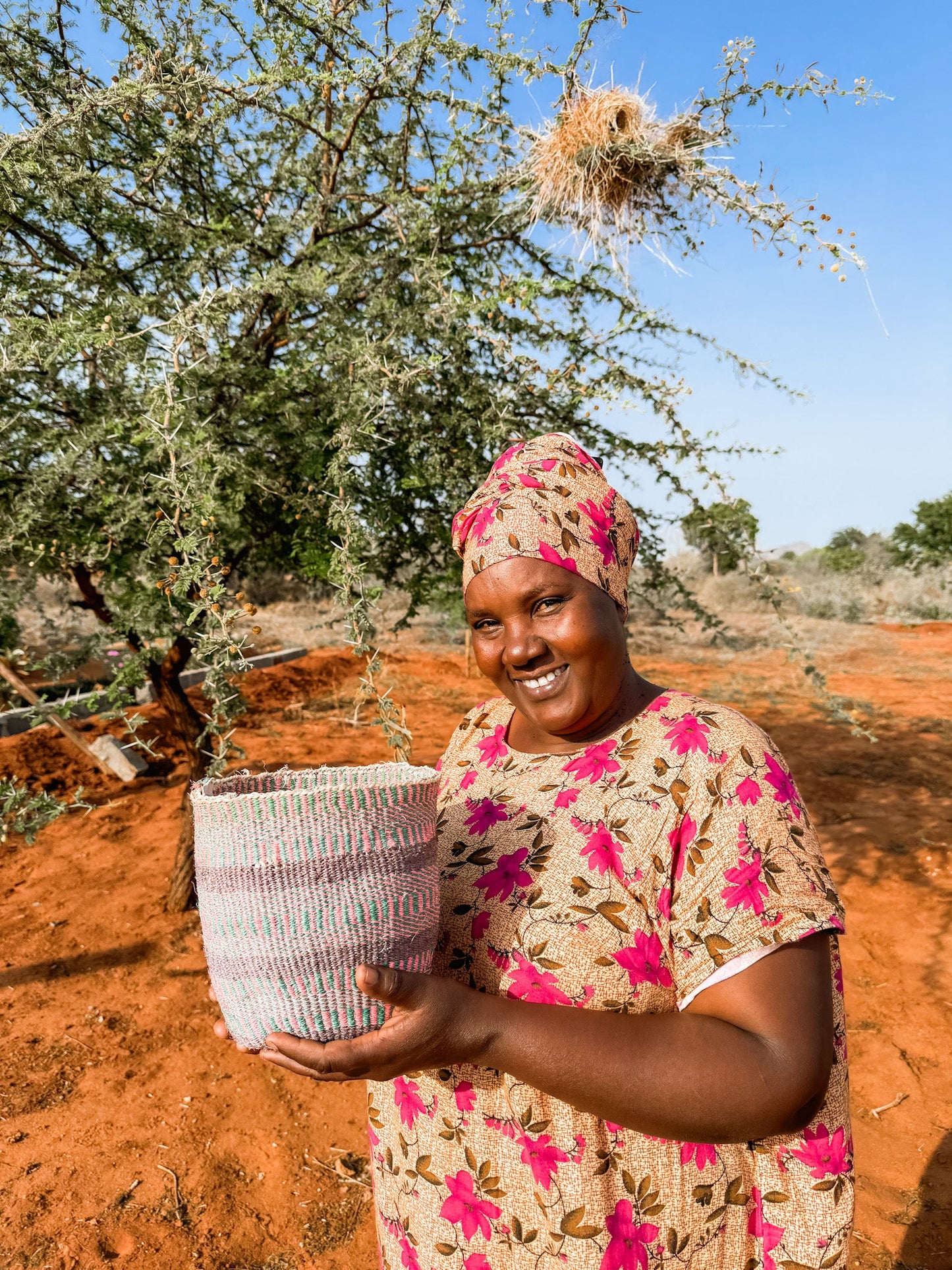 A smiling woman in a floral dress and headscarf stands outdoors on red earth near a leafy tree with a large bird nest, holding the Fine Weave Sisal Basket, fair trade from Kenya, under a clear blue sky.