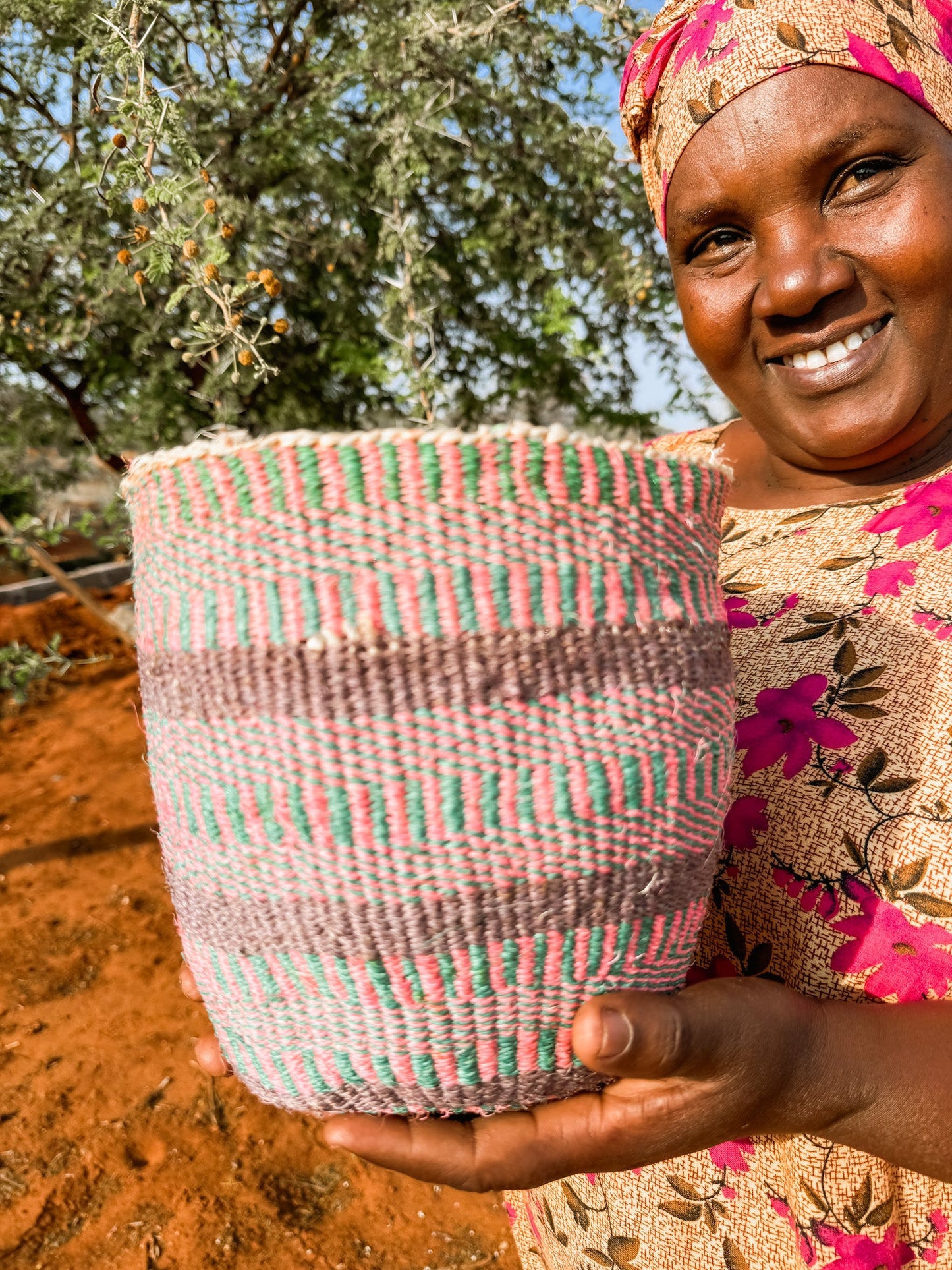 A smiling woman in a colorful dress and headscarf holds the Fine Weave Sisal Basket, featuring pink, green, and beige stripes. She stands outdoors on reddish soil with a tree behind her, showcasing this handcrafted Kenyan basket.