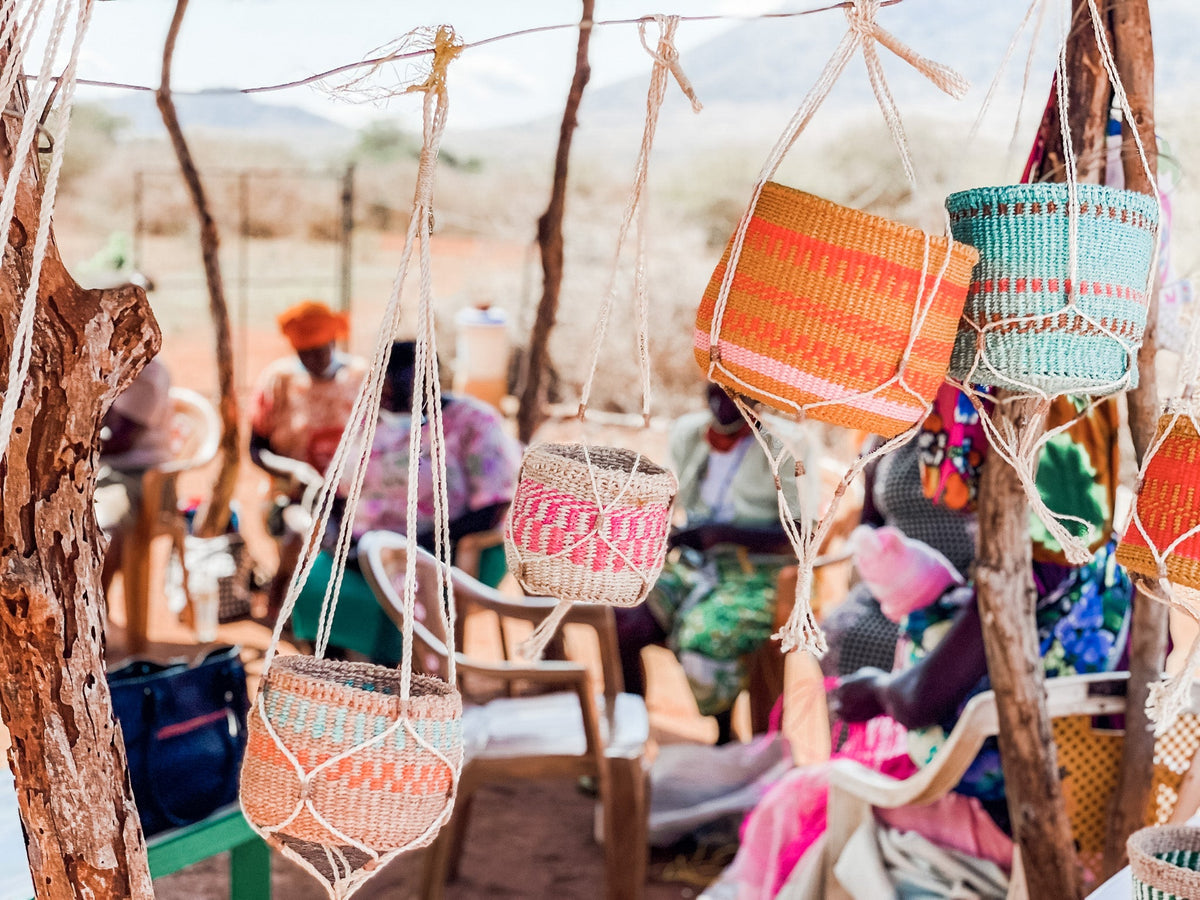 Plant & Basket Hangers are displayed hanging from strings in the foreground, while people in bright outfits gather outdoors amid trees and mountains under sunny skies.