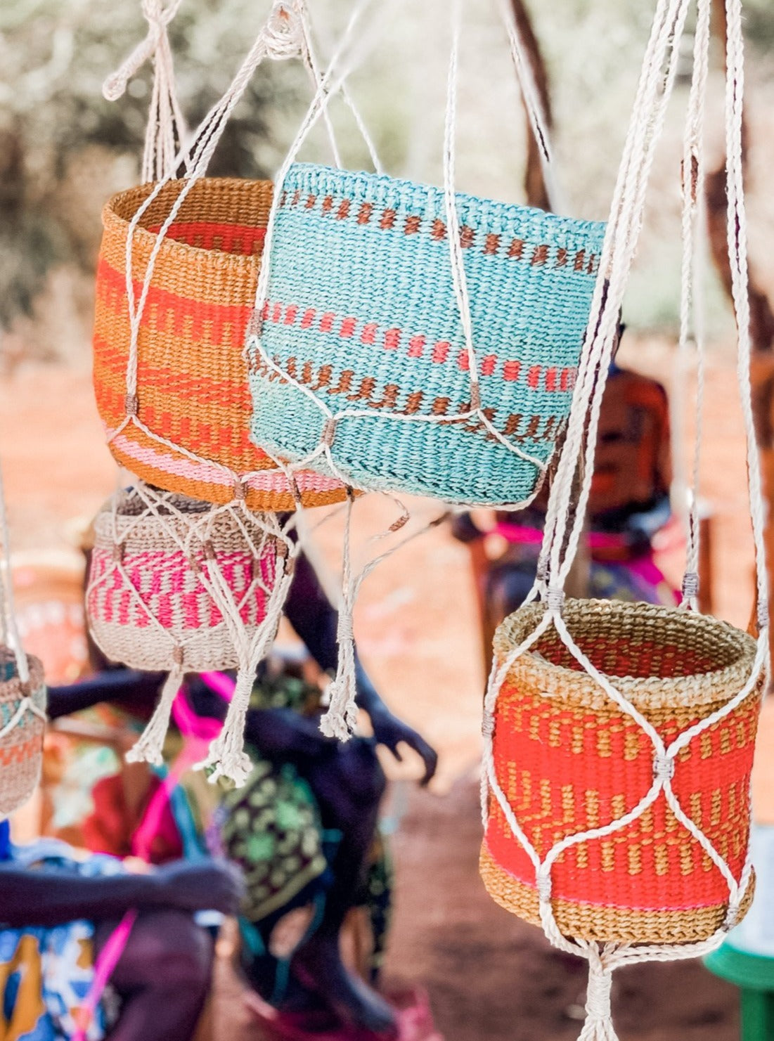 Plant & Basket Hangers display vibrant woven baskets in red, orange, pink, and blue on white macramé hangers. The background features an earthy outdoor scene with people softly blurred.