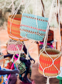 Plant & Basket Hangers display vibrant woven baskets in red, orange, pink, and blue on white macramé hangers. The background features an earthy outdoor scene with people softly blurred.