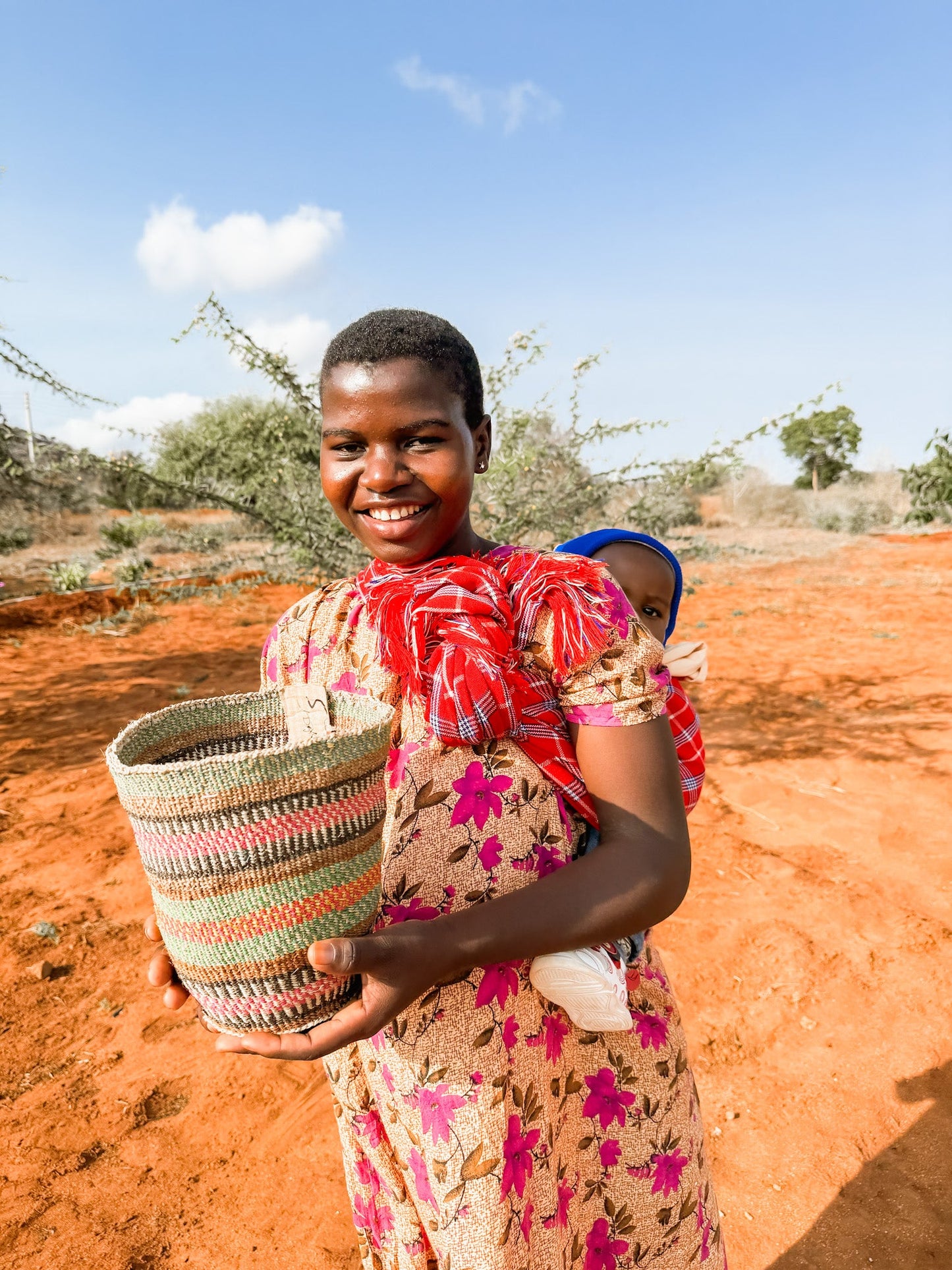 A smiling woman in a patterned dress stands on red soil, holding the Fine Weave Sisal Basket. She carries a baby wrapped in a red cloth on her back, with a clear blue sky and scattered trees behind her.