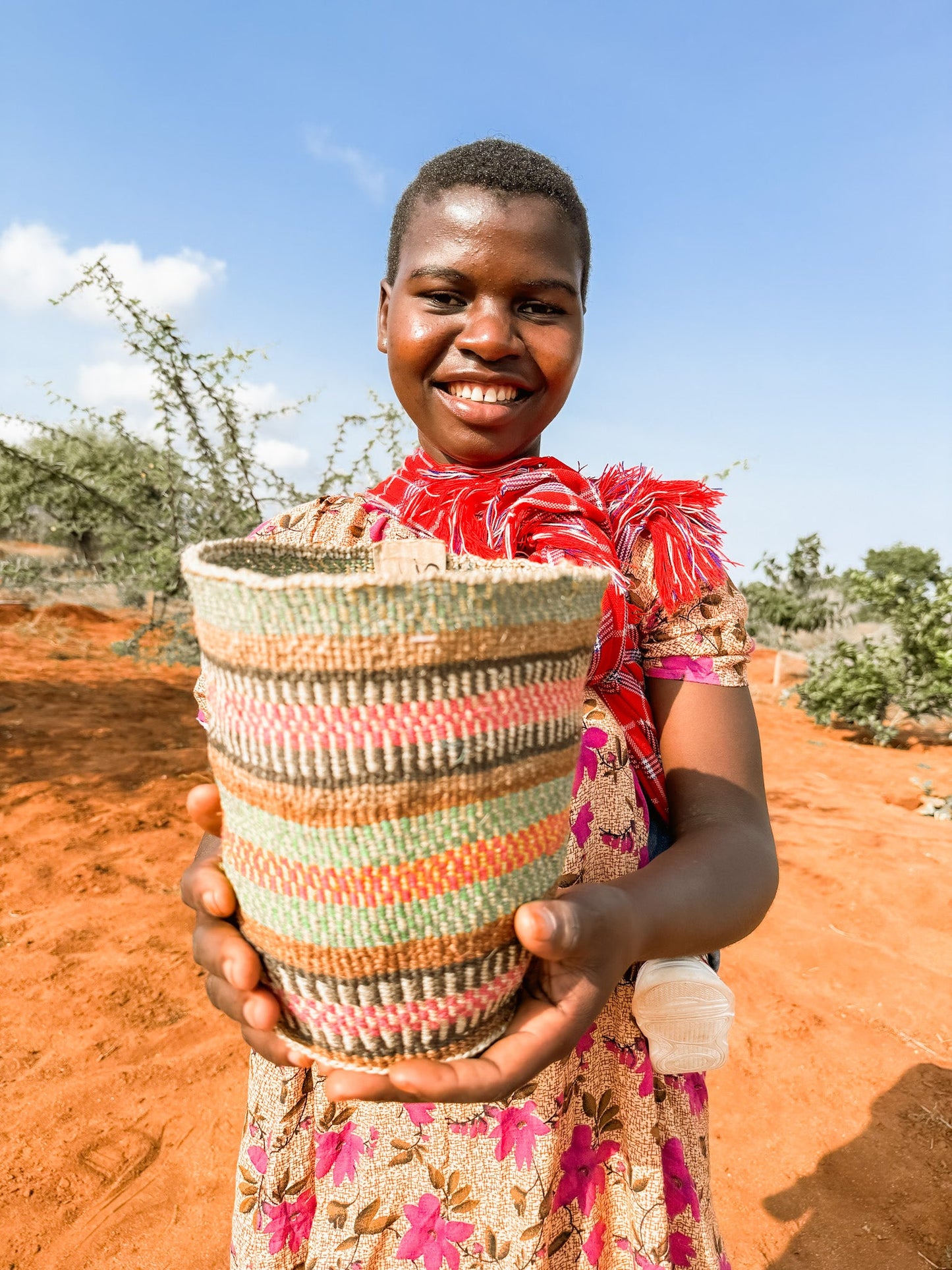 A smiling woman stands outdoors on red soil, holding a Fine Weave Sisal Basket toward the camera. She wears a floral dress and a red decorative shoulder piece, with sparse vegetation under a blue sky in the background.