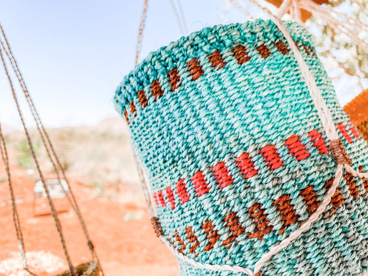 Close-up of a Plant & Basket Hangers woven basket with turquoise, brown, and red patterns, hanging outdoors on a sunny day with blurred orange soil and greenery in the background.