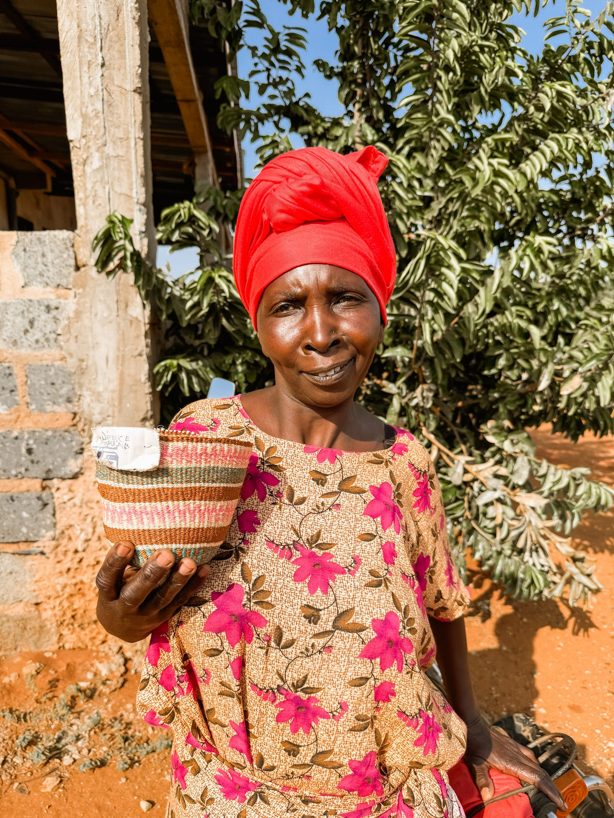 A woman in a bright red headwrap and floral dress stands outdoors, smiling slightly as she holds a Fine Weave Sisal Basket. Trees and a partially built structure are visible in the background.