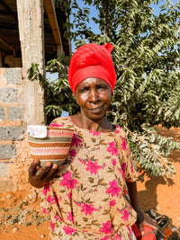 A woman in a bright red headwrap and floral dress stands outdoors, smiling slightly as she holds a Fine Weave Sisal Basket. Trees and a partially built structure are visible in the background.