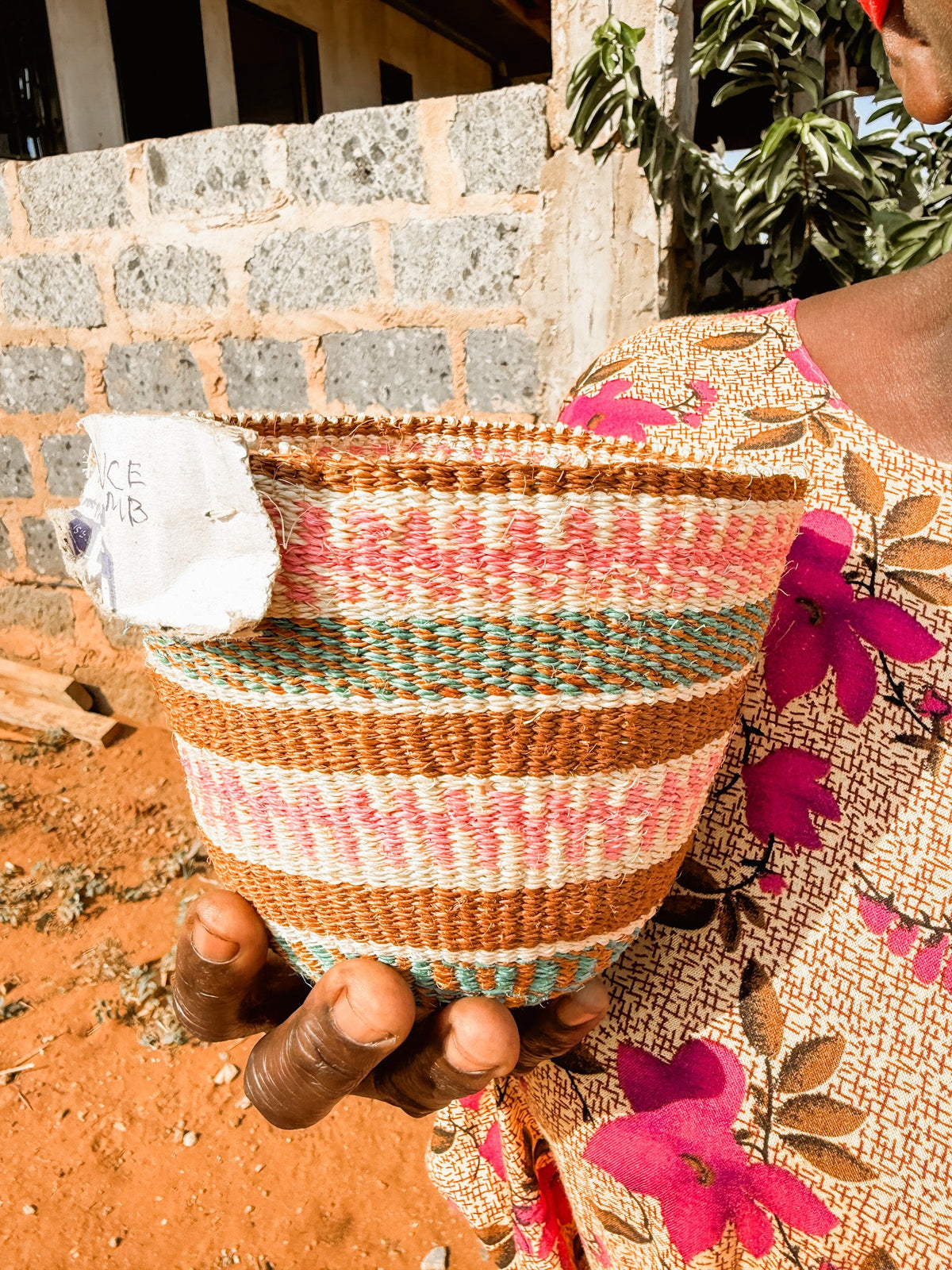 A person in a floral-patterned outfit holds a colorful Fine Weave Sisal Basket with a white price tag, standing outside near a stone wall and reddish-brown soil.